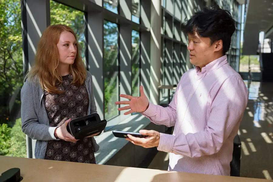 A male professor in a pink business shirt demonstrates a virtual reality headset to a redheaded female student in a sunny lab with big windows.