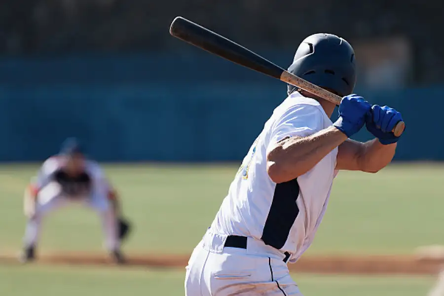 Seen from behind, a baseball player grips his bat as he prepares to swing. Another player in the background crouches dramatically, watching him.