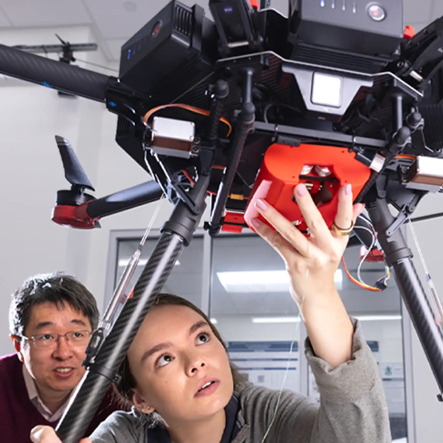 A female student looks up in wonder while working under a large drone, with a male professor watching her intently.
