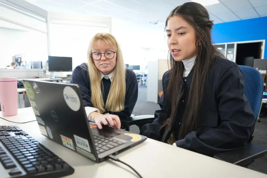 Two female students in a large corporate office look at a computer screen together.