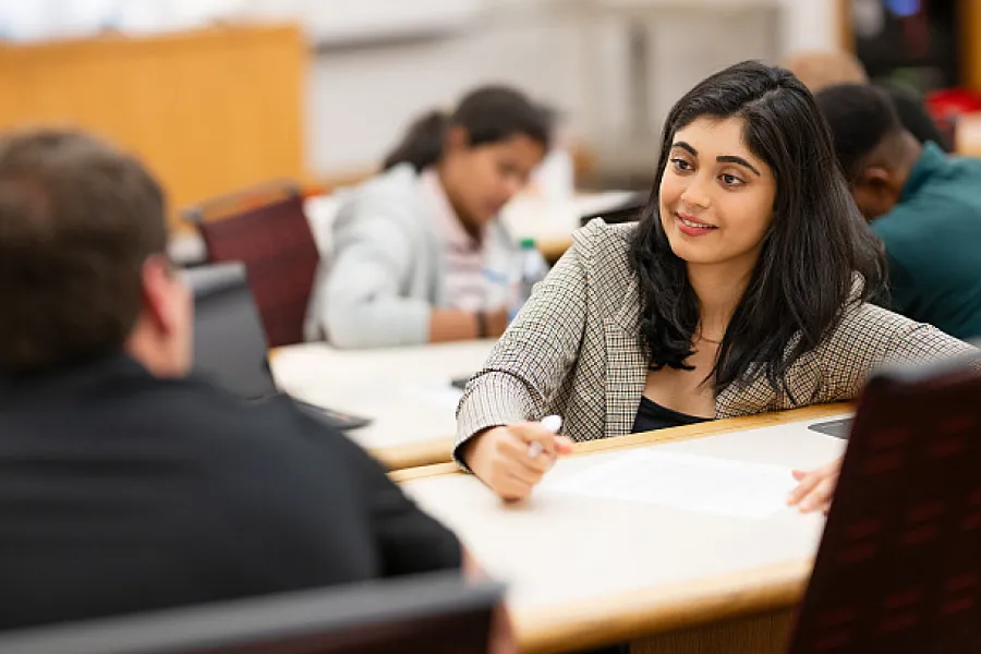 A young woman in a checkered business suit taking notes while conversing with an older male.