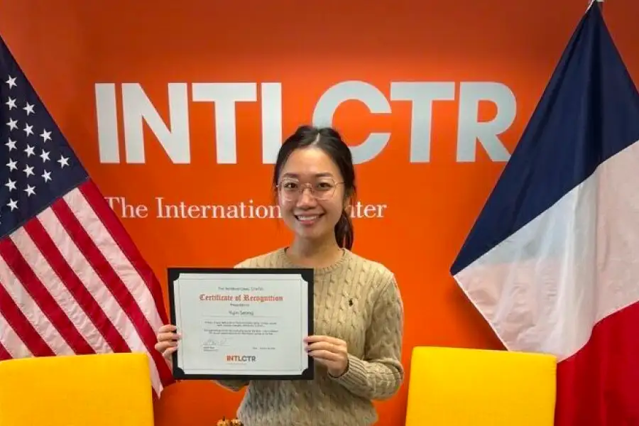A young woman wearing glasses and a tan cable-knit sweater holds a certificate in front of a wall of US and foreign flags.