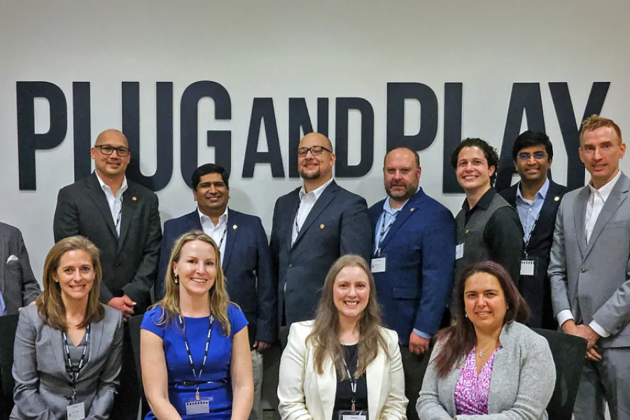 A multi-racial group of male and female businesswomen posing for a group picture in front of a wall graphic, reading in bold type, plug and play.
