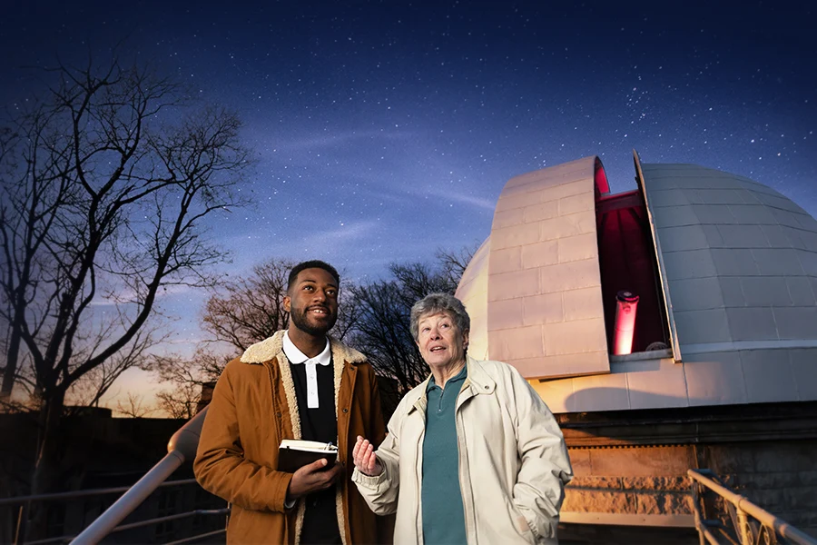 Two people standing in front of an observatory look up, with the night sky illuminating them from behind.