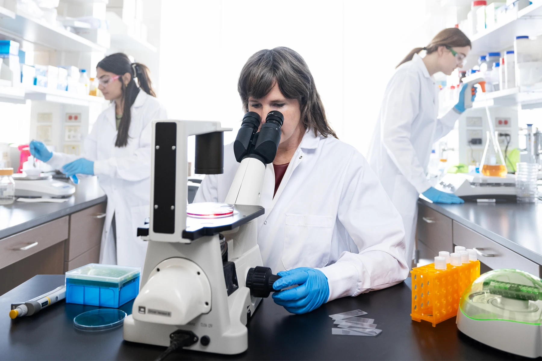 A female scientist looks into a microscope while two student researchers work in the lab behind her.